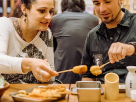 Host sharing grilled skewers in a standing bar.