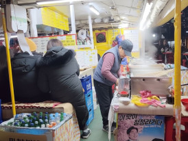 Plastic food tent stall at night with soju bottles and patrons waiting