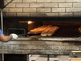 Baker pulling fresh sesame bagels from a wood-fired oven, steaming hot and golden
