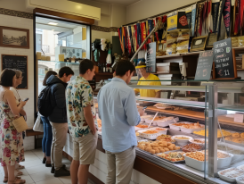 Small deli counter with salami hanging and owner making sandwiches for waiting customers