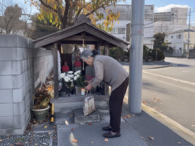 Elderly woman offering flowers at a small roadside shrine in a quiet Tokyo neighborhood