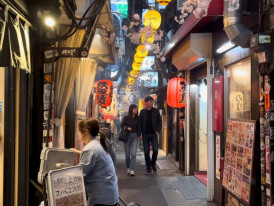 People walking through a narrow Tokyo izakaya alley with lantern-lit restaurants and street food menus in the evening