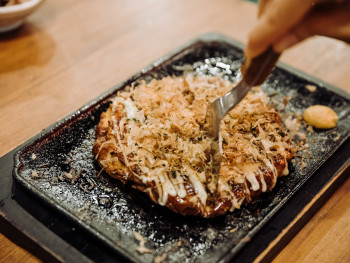 Okonomiyaki being prepared on a hot griddle with chef's hands visible, steam rising from the cooking surface Okonomiyaki being prepared on a hot griddle with chef's hands visible, steam rising from the cooking surface