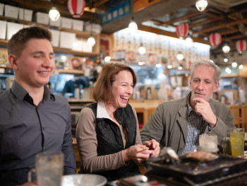 CU host laughing with couple at izakaya counter. CU host laughing with couple at izakaya counter.