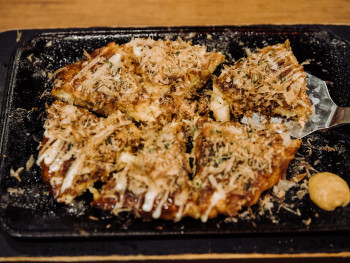 A host demonstrating proper okonomiyaki preparation technique while guests watch and learn A host demonstrating proper okonomiyaki preparation technique while guests watch and learn