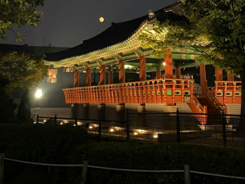 The moon peeking from behind Changdeokgung Palace with its traditional architecture illuminated under the night sky The moon peeking from behind Changdeokgung Palace with its traditional architecture illuminated under the night sky