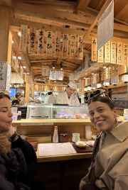 Two women seated at a sushi counter with a chef preparing food in Tokyo.