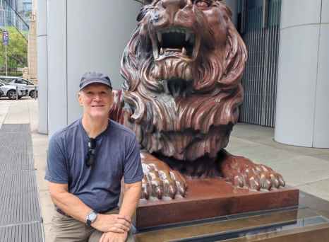 Man seated beside lion statue outside HSBC Main Building, Hong Kong.