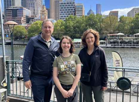 Skyline of Melbourne visible behind three people standing by the river.