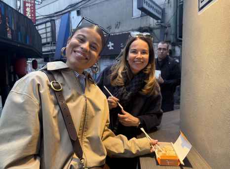 Two women with chopsticks and a food box in a Tokyo alleyway.
