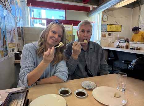 Two people eating at a wooden table with sushi plates and soy sauce.