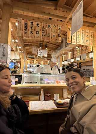 Two women seated at a sushi counter with a chef preparing food in Tokyo.
