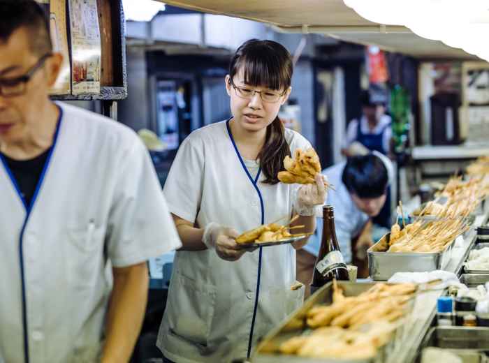 Woman preparing kushikatsu at street stall with neon lights in the background.