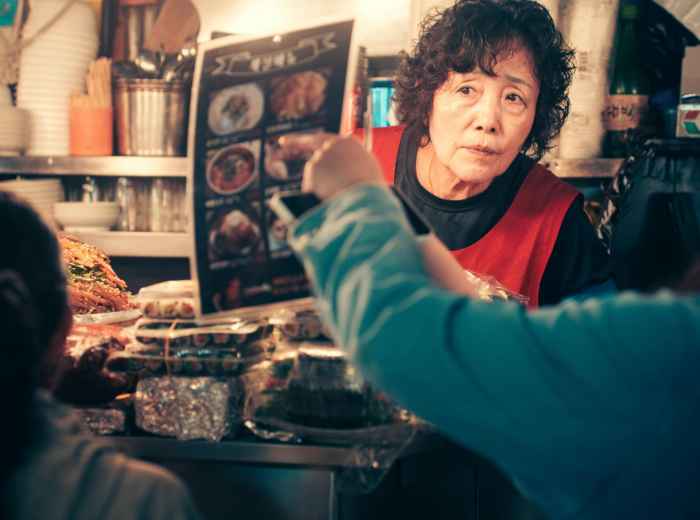 An elderly vendor at a small kushikatsu stall carefully preparing fried skewers while customers wait at the narrow counter Photo by Paul Bill on Unsplash
