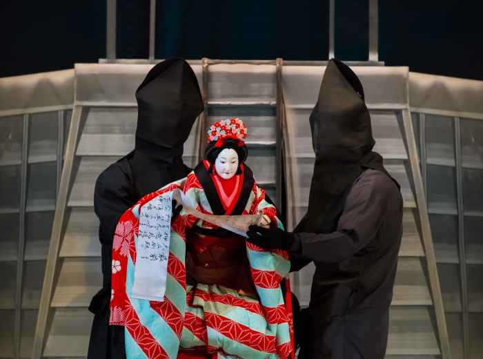 A Bunraku puppeteer in traditional attire performing on stage at the National Bunraku Theatre, with richly detailed puppets and narrators in the background. photo by 177878081 on shutterstock