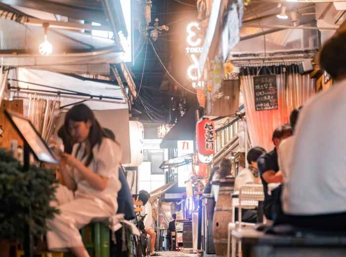  A traveler seated at a modest Osaka eatery, pointing at a handwritten Japanese menu while the chef smiles behind the counter; residential streets and laundry-draped balconies visible through the window. Photo by Satoshi Hirayama on pexels