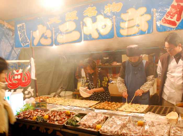 Colorful Osaka street food market with smoke rising from takoyaki stalls. Image by chou_i_ci from Wiki Commons