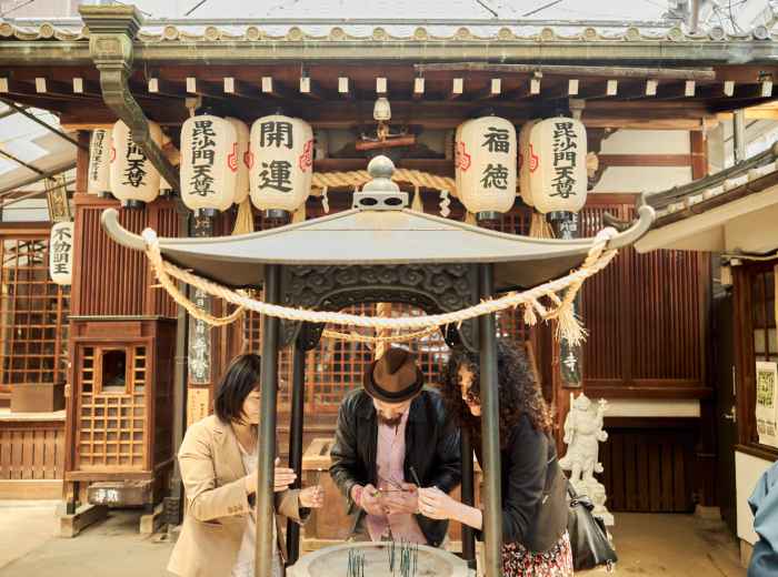 A small group of travelers discovering a tucked-away shrine hidden in a quiet Osaka alley, surrounded by stone lanterns and lush greenery. 