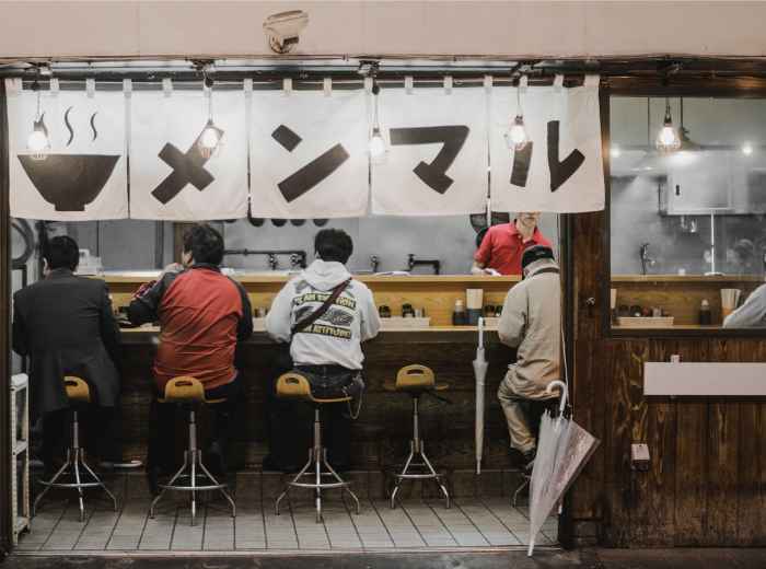 Late-night ramen stand with neon lighting and a few customers eating at the counter.