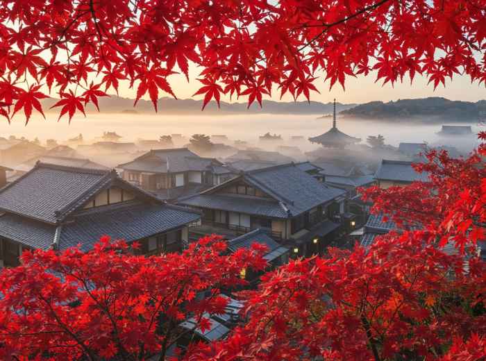 Morning mist rising over traditional Kyoto rooftops with red maple leaves in foreground.
