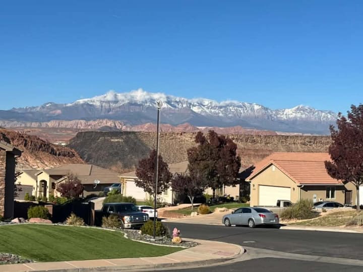 A quiet neighborhood street of single-story homes, with the red cliffs behind them and the snow-capped Pine Valley Mountains beyond, under a clear blue sky.