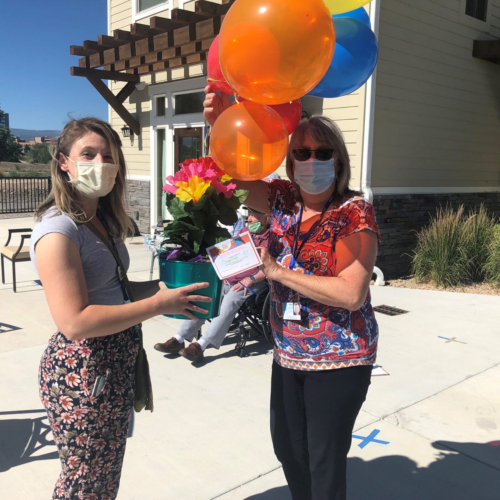 Two people in face masks handing over a potted flower arrangement with Day of Joy balloons