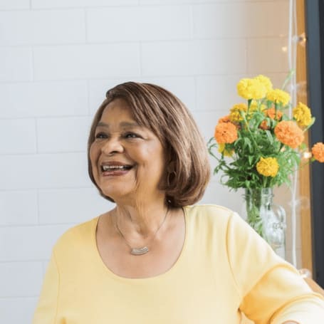 A smiling senior woman in a yellow cardigan seated beside a vase of orange flowers
