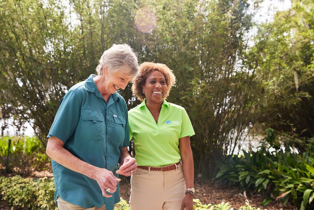A caregiver walking outdoors through a garden with a senior woman
