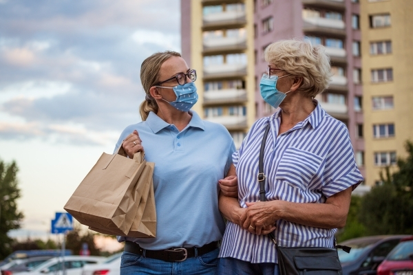 Caregiver wearing a surgical mask and holding two grocery bags walking arm-in-arm with older woman wearing a surgical mask.