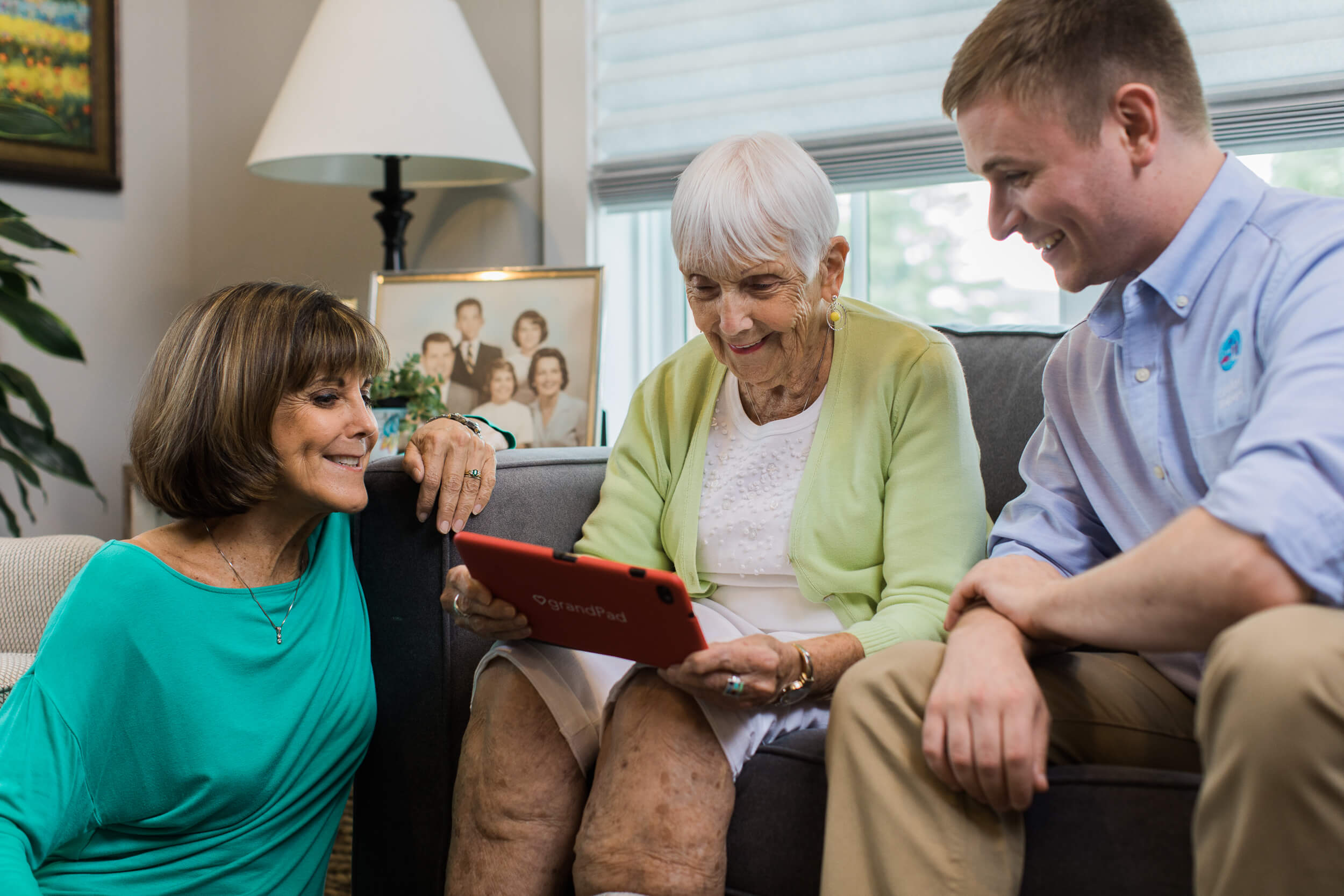 Photo of senior holding a tablet with her daughter and caregiver sitting next to her in the living room