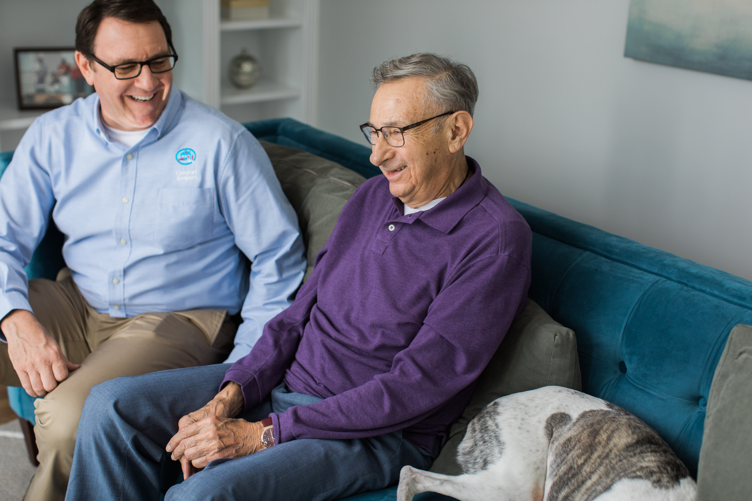 Comfort Keepers caregiver and older adult male sitting on sofa with a sleeping dog next to man.