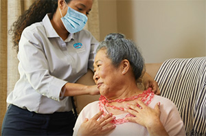 Caregiver with mask helping senior put on beaded necklace