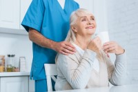 Caregiver standing behind senior seated at kitchen table drinking tea