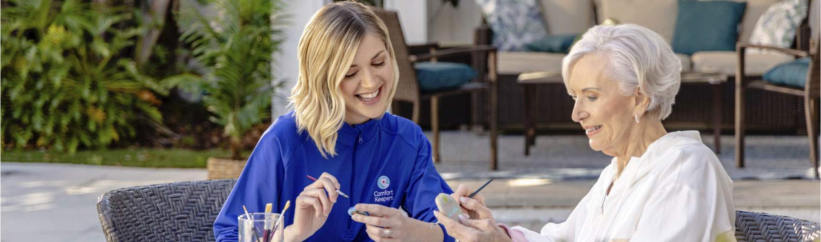 Caregiver and client sit outside on a shady patio, painting, and laughing