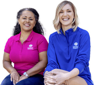 Two caregivers sit side by side smiling one in a pink Comfort Keeper polo and the other in a blue Comfort Keeper pullover