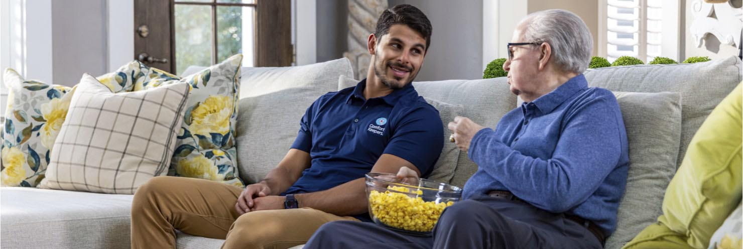 Caregiver and client smiling and eating popcorn sitting on a couch in living room