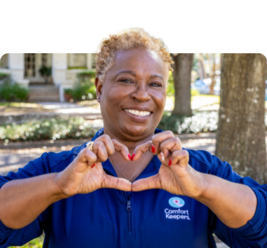 Caregiver in dark blue Comfort Keeper company polo making a heart shape with their hands as they smile and pose for a photo.