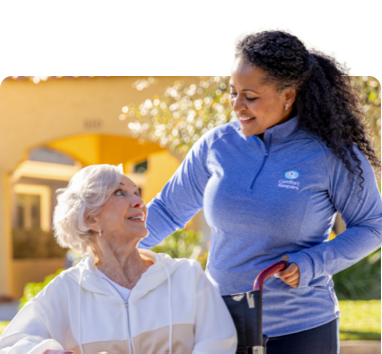 Caregiver pushing client in wheelchair as they smile warmly at one another enjoying a stroll outside on a sunny day.