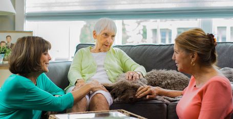 Elderly lady is sitting on the couch conversing with two ladies sitting on the floor in front of her.
