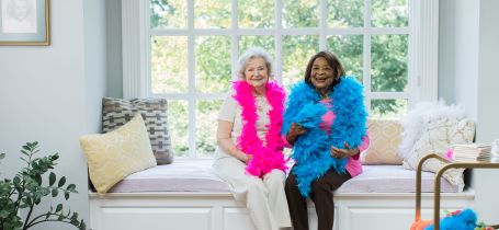 Two senior ladies sitting in window seat with smiling faces and feather boas on