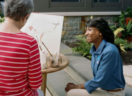 Photo of senior painting on a canvas while a caregiver keeps her company