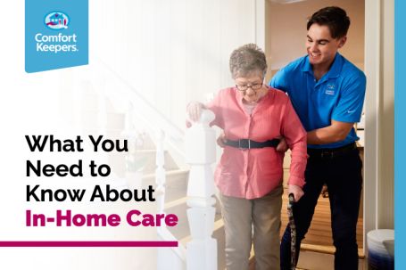 An elderly woman is being assisted walking through a home a younger man from comfort keepers. She is holding onto a white staircase railing with one hand and holding a cane in the other. The younger man is supporting her at the waist. Caption reads "What You Need to Know About In-Home Care"