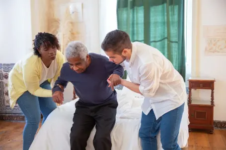 Two caregivers assisting a senior get out of bed and stand up