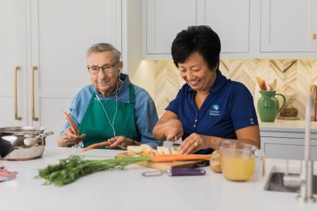 A woman with stands next to and elderly man at a kitchen island. She is cutting vegetables and smiling. He is wearing wired earbuds and drumming on the island using the carrot sticks. 