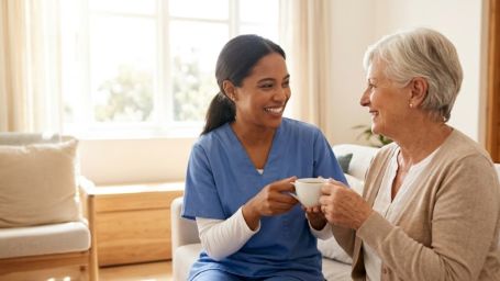 A caregiver and senior share a warm, joyful moment with tea in a sunny living room.