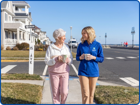 Comfort Keepers caregiver and a senior woman walking and sharing coffee along a shore street in Belmar, NJ.