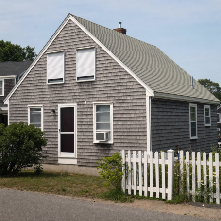Weathered gray shingled Maine Cape house with closed shades and window AC unit on a hazy August afternoon.