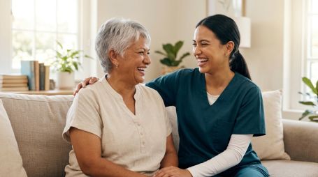 A senior woman and her caregiver laugh together in a sunlit living room.