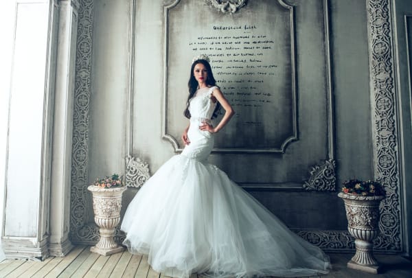 A woman in a stunning white mermaid-style wedding gown stands in an ornate, vintage-inspired room with intricate architectural details and decorative elements.