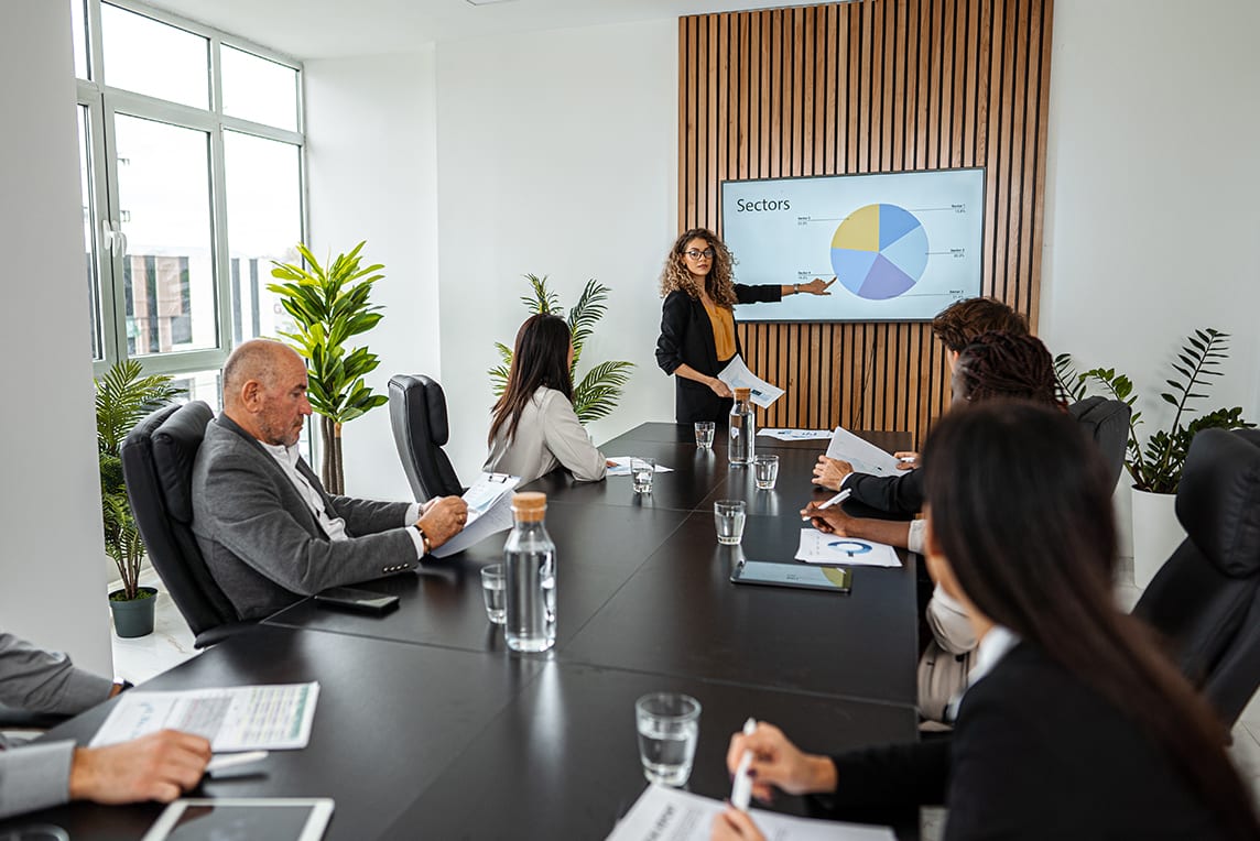 Employees gathered around conference room table, watching presentation on TV